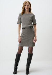 Robe grise à motifs avec des manches courtes, silhouette ajustée et boutons décoratifs à la taille ; portée avec des bottes noires jusqu'au genou.