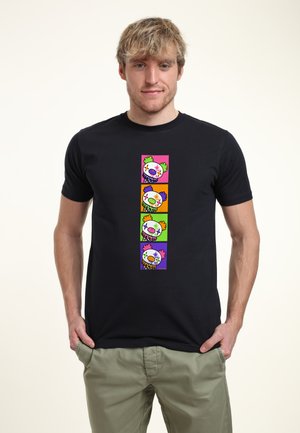 Ung mand iført en sort t-shirt med en lodret række af fire farverige klovneansigter i firkantede rammer, poserer med hænder i lommerne.