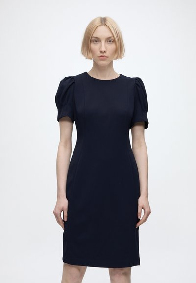 Femme blonde portant une robe bleu marine cintrée avec des manches courtes bouffantes, debout devant un fond clair uni, face à nous.