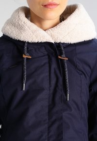 Námorně modrá bunda s krémovou fleece podšívkou na kapuci, se stříbrnými prvky a pletenými šňůrkami. Texturovaná tkanina s kapsami.