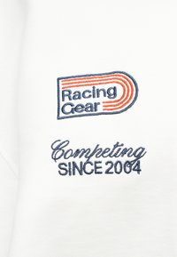 Bílá látka s vyšitým textem „Racing Gear“ a „Competing Since 2004“, zdobená oranžovými a modrými liniemi tvořícími stylizovaný tvar.