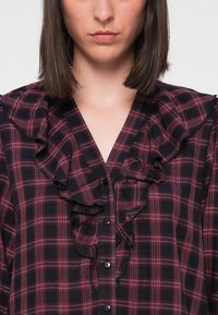 Black and red plaid shirt featuring ruffled front detail, button closure, and a V-neckline. The fabric has a textured appearance.