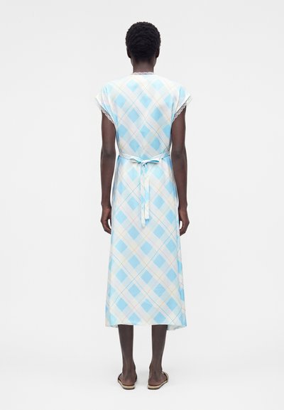 Γυναίκα που φαίνεται από πίσω φοράει ένα μπλε και λευκό μίντι φόρεμα με μοτίβο διαμαντιού, κοντά μανίκια και ζώνη στη μέση, σε συνδυασμό με φλατ σανδάλια.