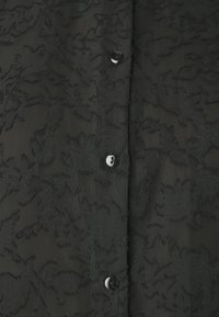 Μαύρη μπλούζα με υφή λουλουδιών από δαντέλα, με κουμπιά μπροστά, μαλακό ύφασμα και διάφανη εμφάνιση. Διακοσμημένη με μαύρα κουμπιά.