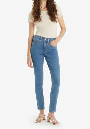 Femme portant un jean skinny bleu taille haute, un haut fitted à manches courtes écru et des sandales blanches à talons blocs et bouts ouverts, debout avec une main dans la poche.