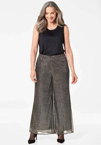 Metallic plissierte Hosen mit weitem Bein und hoher Taille, kombiniert mit einem schwarzen ärmellosen Oberteil. Der Stoff zeigt ein glänzendes, strukturiertes Muster.