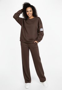 Brązowa bluza o luźnym kroju, z prążkowanym dekoltem i kieszenią na szwie bocznym. W zestawie z dopasowanymi brązowymi spodniami o prostych nogawkach. Białe buty.