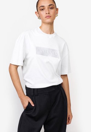 Kobieta w białej koszulce z logo "LAMANIA" i czarnych spodniach z wysokim stanem, pozująca z jedną ręką w kieszeni na białym tle.