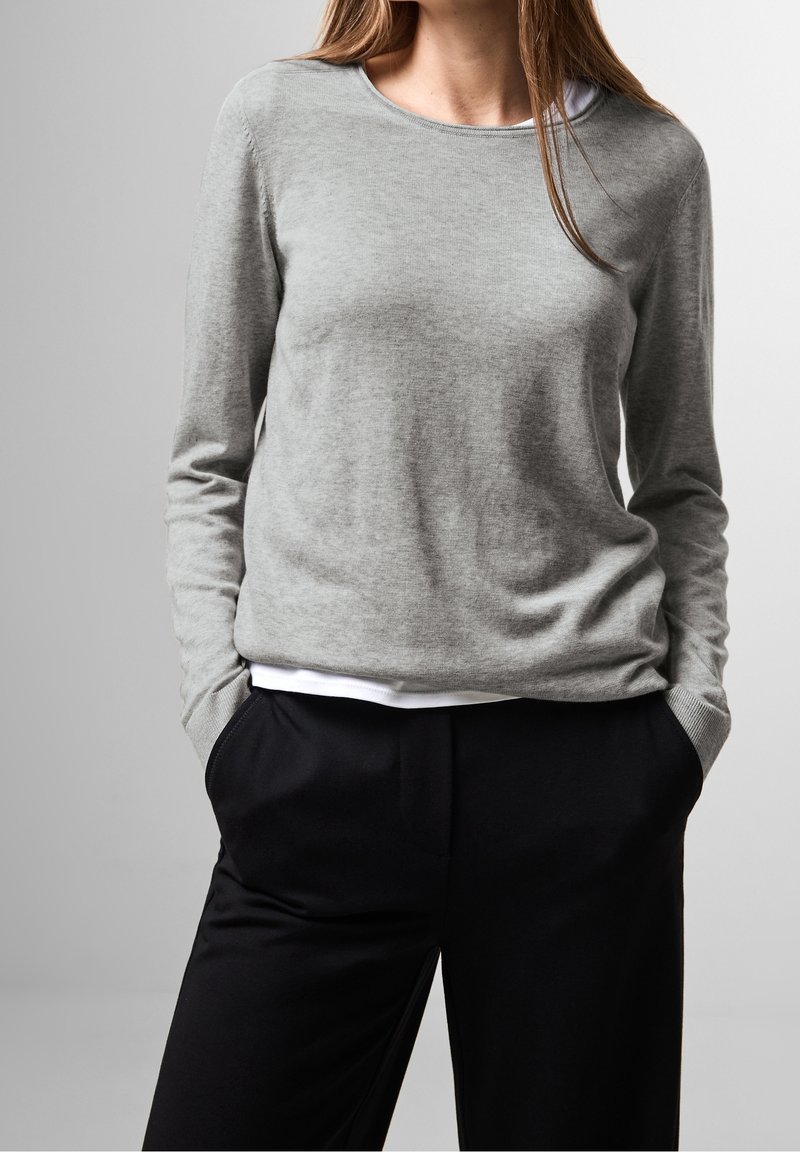 Pull gris à manches longues en tissu tricoté doux, avec un col rond et une coupe décontractée, porté par-dessus une chemise blanche et un pantalon noir.