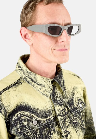 Grijze, rechthoekige zonnebril met donkere lenzen. Draagt een gele jas met een zwart printpatroon en een gestructureerde kraag.