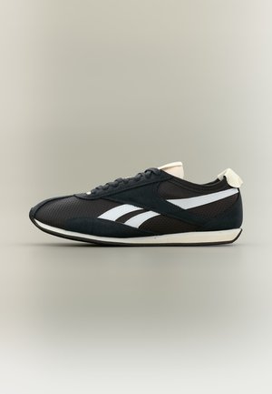 Chaussure de sport noire avec des bandes blanches sur les côtés, des lacets noirs et une semelle blanche sur un fond neutre, vue de profil.