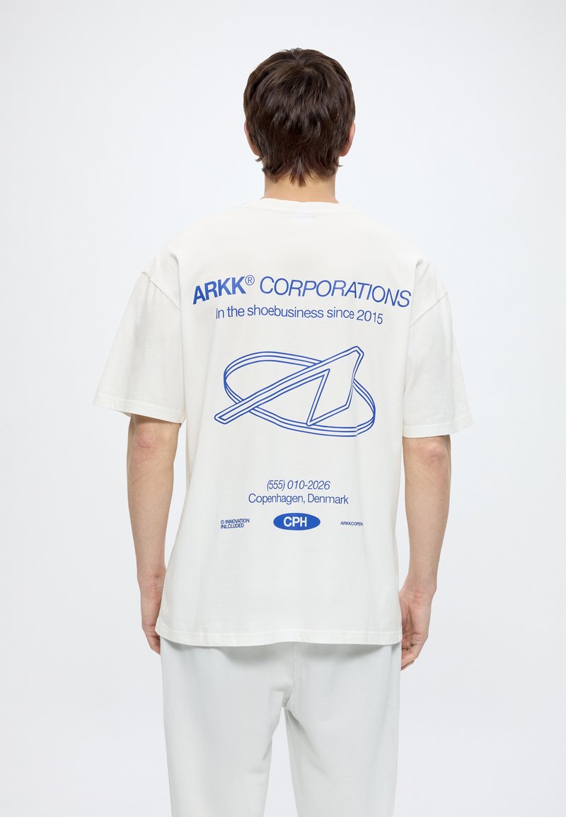 Άτομο που φοράει λευκό T-shirt με μπλε λογότυπο ARKK Corporations, σύνθημα και στοιχεία επικοινωνίας Κοπεγχάγης στην πλάτη, στέκεται μπροστά σε λευκό φόντο.