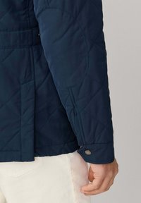 Marineblauwe gewatteerde jas met een gestructureerde stof, elleboogpatches en drukknoopsluiting op de manchetten. Gedetailleerd stikwerk is zichtbaar op het lichaam.