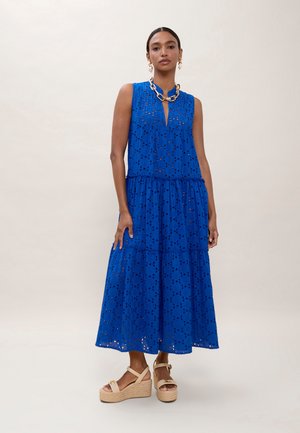 Mujer con vestido midi azul sin mangas con ojales, sandalias cuña beige, collar de cadena dorada y pendientes de perlas sobre un fondo liso.