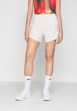 Personne portant un short de sport blanc, des chaussettes Nike blanches, des baskets blanches et un haut court rouge-orange sur un fond uni.