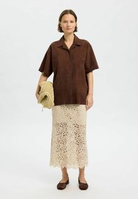 Brun suedeskjorte med korte ærmer, beige hæklede midiskirt med blonder, holder en vævet taske, parret med mørkebrune sko.