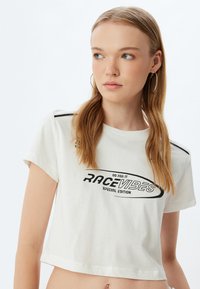 Witte cropped t-shirt met korte mouwen, zwarte grafische tekst "RACE VIBES" en accenten, met een ronde halslijn en zacht katoenmateriaal.