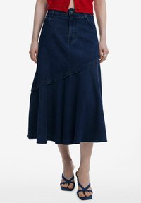 Denimhame, joka on korkea vyötärö ja tumman värinen, epäsymmetrinen helma ja virtaava siluetti. Viimeistelty tikkausviivoilla ja edessä olevalla napilla.