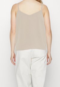 Mujer vestida con una blusa sin mangas beige de tirantes finos y pantalones blancos holgados, mostrada de espaldas contra un fondo blanco liso.