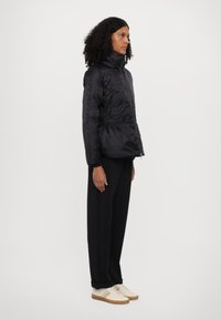 Black quilted jacket with high collar and zipper, featuring a textured pattern. Worn with black pants and beige sneakers with colored stripes.