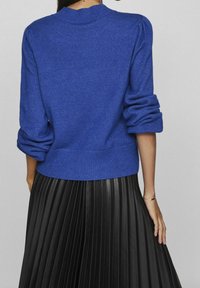 Maglione blu lavorato a maglia con colletto a coste e maniche a sbuffo, abbinato a una gonna nera plissettata. La texture sembra morbida e di peso medio.
