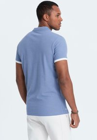 Modrá polo košile s krátkým rukávem a bílým lemem na límci a rukávech. Hladká tkanina s přiléhavým střihem, zobrazeno zezadu.