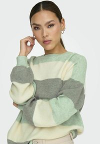Pull rayé vert menthe et gris avec un col rond, texture côtelée et poignets élastiques; porté avec un pantalon beige neutre.