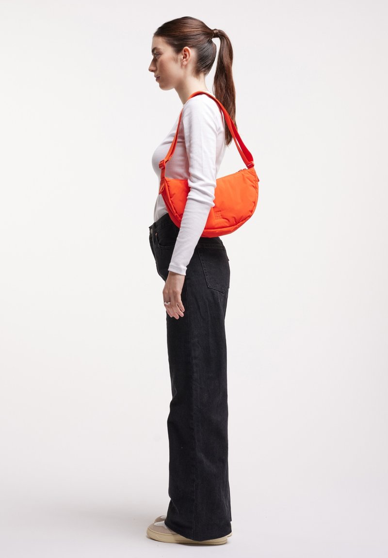 Femme avec queue de cheval brune, de profil vers la gauche, portant une chemise blanche à manches longues, un pantalon noir, des chaussures beiges, et portant un sac à bandoulière orange vif.