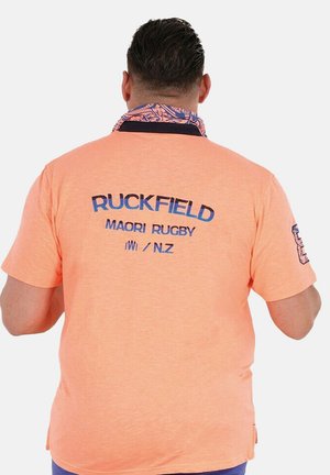 Homme aux cheveux courts et foncés portant un polo pêche avec l'inscription bleue "Ruckfield Maori Rugby N.Z" au dos et un col à motifs, vu de dos.