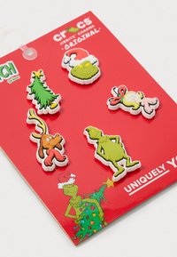 Breloki Crocs Jibbitz na czerwonej karcie; wzory obejmują zielonego Grincha, choinkę oraz psa z rogami, z różnymi kolorami i detalami.
