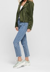 Giacca da biker in suede verde oliva con zip argento, abbinata a jeans azzurri a vita alta e sneakers verde chiaro. Look casual.