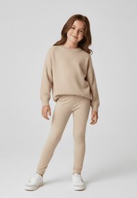 Beżowy sweter na drutach z opuszczanymi ramionami, połączony z jasnobeżowymi legginsami i białymi trampkami. Strój ma luźny krój i gładką fakturę.