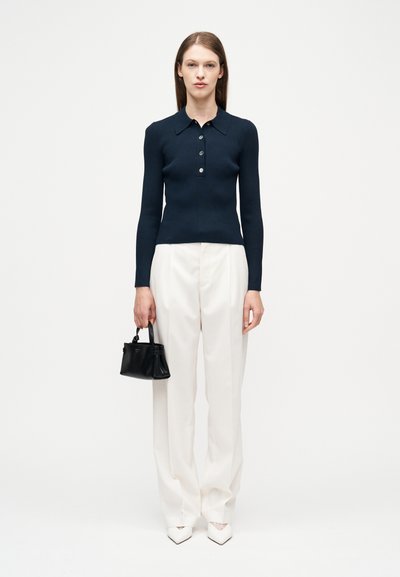 Γυναίκα που φοράει μπλε μακρυμάνικο πόλο, λευκό παντελόνι φαρδύ, λευκά μυτερά παπούτσια, κρατώντας μια μικρή μαύρη τσάντα, στέκεται σε λευκό φόντο.