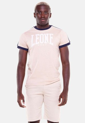Giovane uomo con capelli biondi corti che indossa una maglietta beige chiaro "LEONE 1947" con bordatura blu navy e pantaloncini abbinati, in piedi su sfondo bianco.