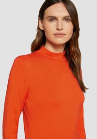 Langärmliges, hochgeschlossenes Oberteil aus leuchtend orangefarbenem Stoff. Glatte Textur mit taillierter Silhouette, keine sichtbaren Muster oder Beschläge.