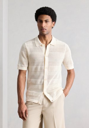 Άνδρας που στέκεται με το ένα χέρι στην τσέπη, φοράει μπεζ πουκαμίσα πλεκτή με κοντά μανίκια, κουμπιά και γιακά, καθώς και μπεζ παντελόνι.