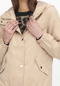 Woman wearing beige hooded jacket over black and beige patterned sweater, fastening jacket zipper and holding collar.