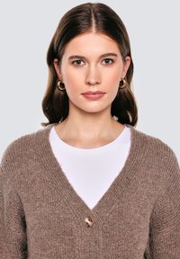 Braune Strickjacke mit V-Ausschnitt, ausgestattet mit einem einzelnen runden Knopf. Getragen über einem weißen Hemd, zeigt einen strukturierten Stoff.