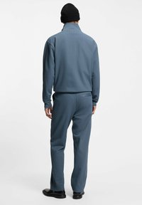 Man facing away wearing a blue pinstripe suit with a high collar, black beanie, and black shoes against a plain white background.