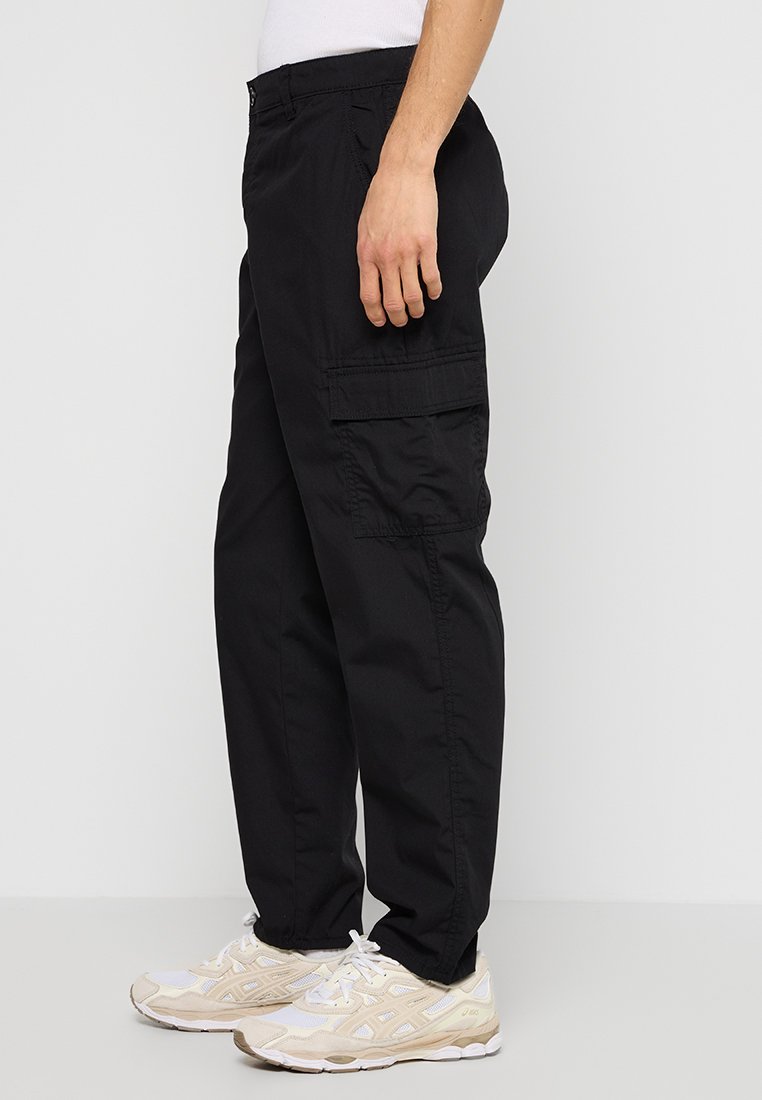 Pantalon cargo noir avec de multiples poches latérales, fabriqué en tissu durable. Il présente une coupe décontractée et une taille à cordon. Porté avec des baskets blanches.