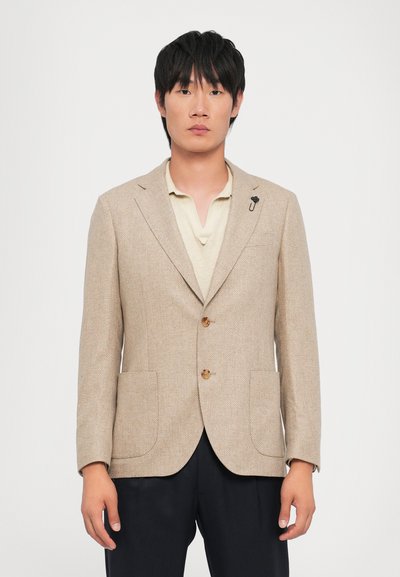 Blazer beige en chevrons avec deux poches avant, boutons en bois et une petite broche d'accent. Porté sur un polo crème, associé à un pantalon sombre.