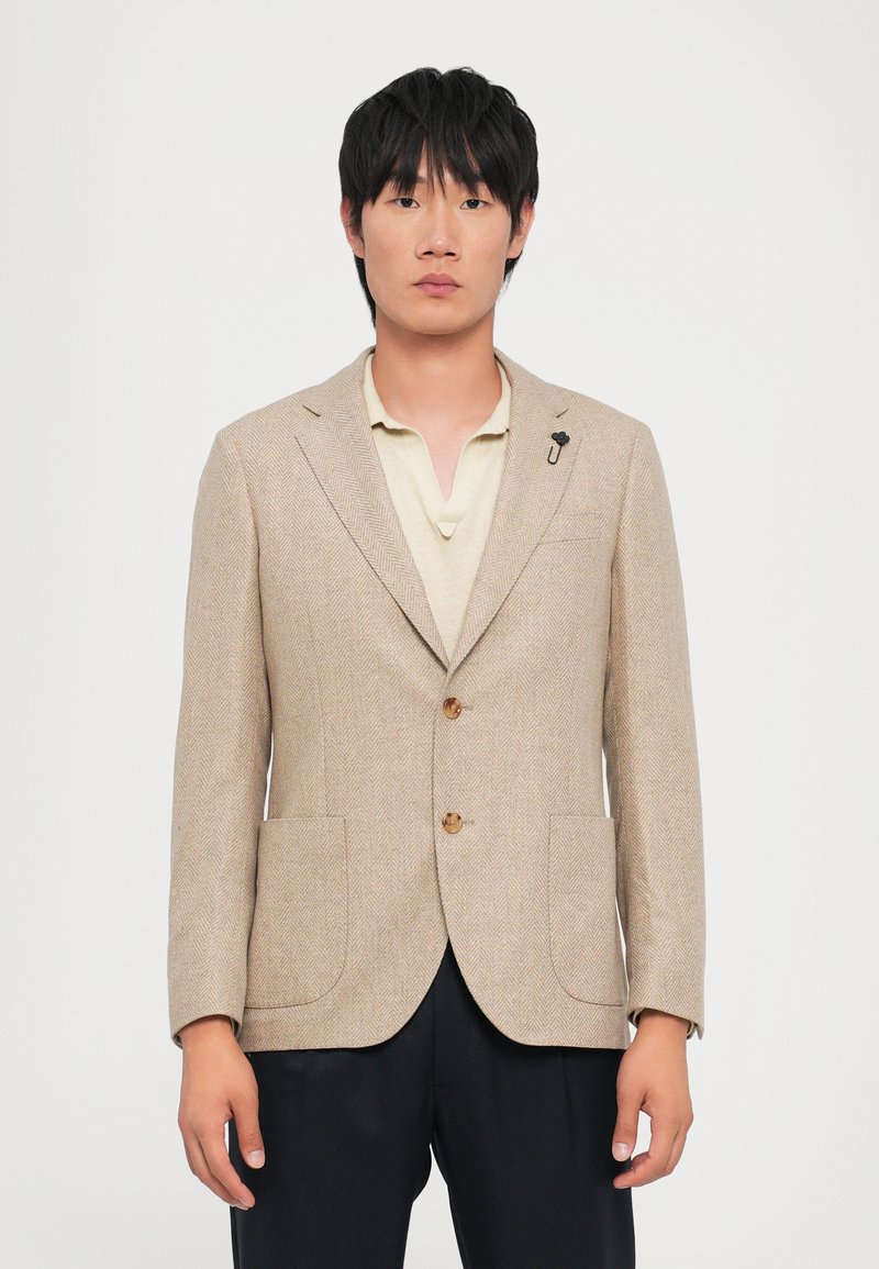 Beige Fischgrätblazer mit zwei vorderen Taschen, Holzknöpfen und einem kleinen Akzent-Pin. Getragen über einem cremfarbenen Poloshirt, kombiniert mit dunklen Hosen.