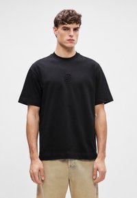 Μαύρο βαμβακερό T-shirt με στρογγυλή λαιμόκοψη, κοντά μανίκια και ανεβασμένο λογότυπο "B" στο στήθος. Το ύφασμα είναι λείο και απλό.