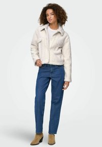 Donna con capelli ricci, che indossa una giacca di pile bianca, jeans blu e stivaletti in camoscio beige, in piedi contro uno sfondo chiaro uniforme.