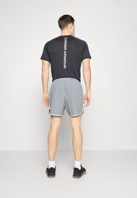 Man som står med ryggen mot kameran, iförd en svart Under Armour-t-shirt, ljusgrå shorts, vita strumpor och svarta träningsskor.