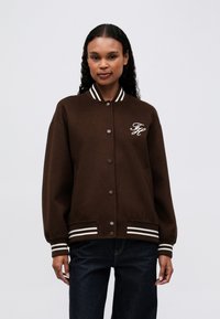 Mujer que lleva una chaqueta varsity marrón con botones, con puños y dobladillo de canalé de rayas blancas, y con iniciales bordadas en blanco en el pecho.
