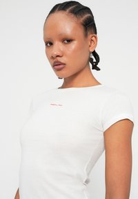 Tricou alb, ajustat, din bumbac moale, cu un mic logo roșu "REPLAY" pe piept, cu decolteu rotund și mâneci scurte.