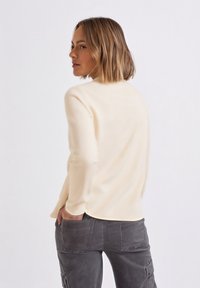 Kermanvärinen resortribillinen pitkähihainen paita pyöreällä pääntiellä, yhdistetty harmaisiin työhaalarihousuihin, joissa on suuret takataskut. Tasainen, istuva materiaali.