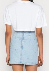 Person som bär en vit kortärmad t-shirt och en ljusblå denim minikjol med en synlig bakdragkedja och subtila broderade mönster.