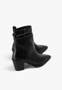 Botas de piel negra hasta el tobillo con punta afilada, tacón bajo en bloque y tachuelas plateadas a lo largo de las costuras laterales. Cierre de cremallera en la parte posterior.